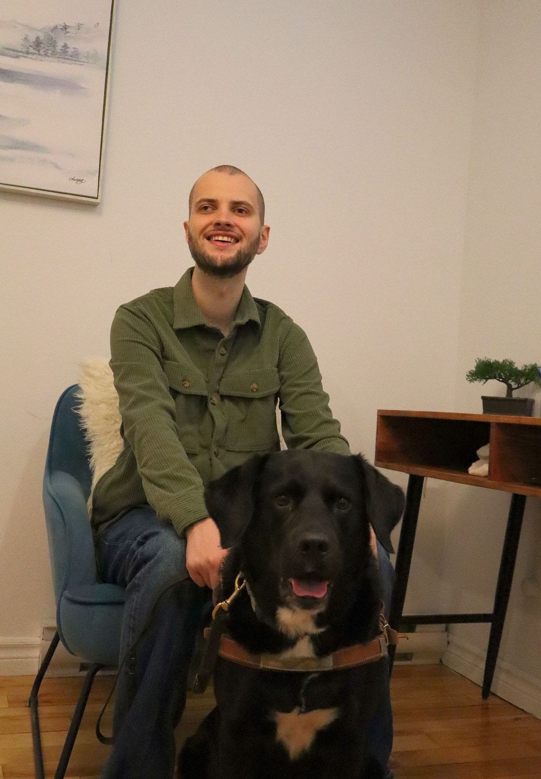 Marc de Montlivault rit, assis dans un fauteuil bleu de son cabinet, une main posée sur son chien-guide, un grand chien noir au harnais de cuir brun.
